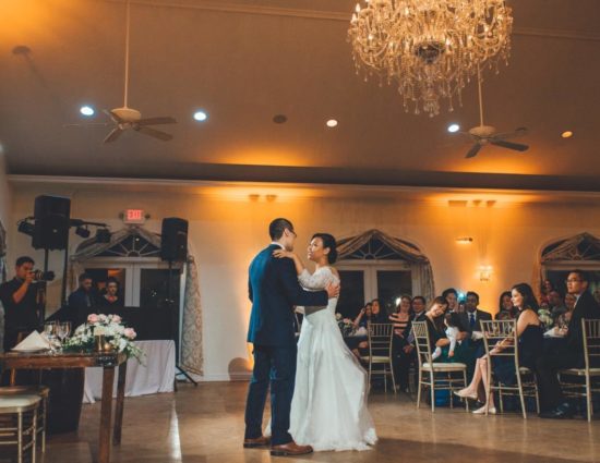 Bride in white dress and groom in dark blue suit enjoying their first dance while people watch