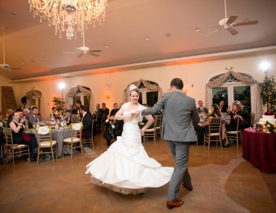 Bride in white dress and groom in gray suit enjoying their first dance while people watch