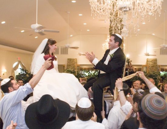 Bride in white dress and groom in black suit sitting on chairs raised in the air with many people celebrating around them