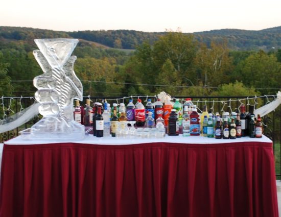 Large table on edge of patio with an ice sculpture, soda, juice, alcohol, and glasses