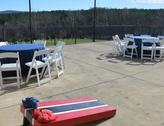 Large concrete patio with two round tables with dark blue tablecloths, white chairs, and a red and blue cornhole board