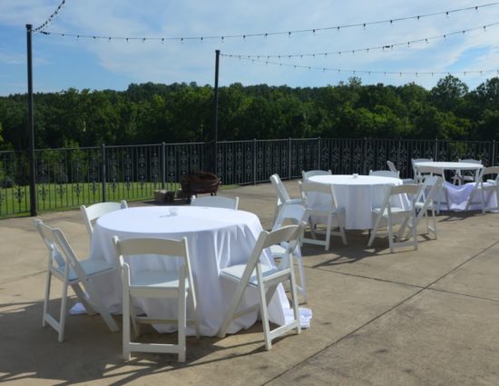Large concrete patio with three tables and chairs with white tablecloths and large trees in the background