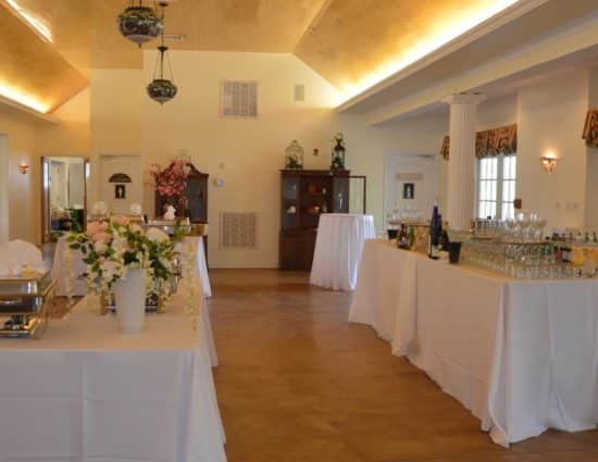 Large room with concrete flooring set up for a wedding reception with white tablecloths, drink station, and buffet station