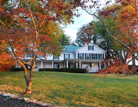 Exterior view of property painted white with green roof and shutters surrounded by large green lawn and trees with fall color