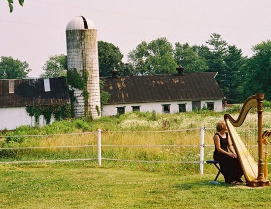 Harp player with black dress sitting near harp with old barn and silo in background