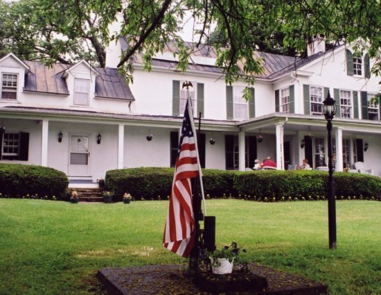 Exterior view of property painted white with green shutters surrounded by green grass and shrubs with American flag out front