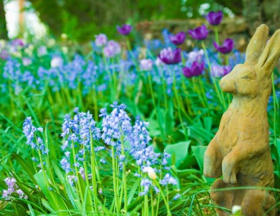 Tan rabbit sculpture sitting in green grasses with blue and purple flowers