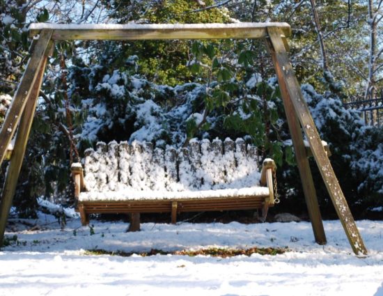 Wooden swing with evergreen trees behind it all covered in snow