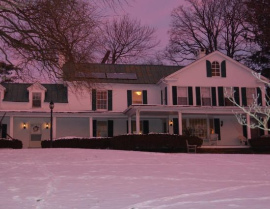 Front of main house painted white with green shutters covered in snow with a light pink hue due to the sun setting