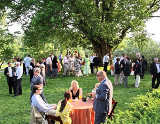 Wedding reception set up on large green lawn near large tree with people sitting at tables and standing around talking