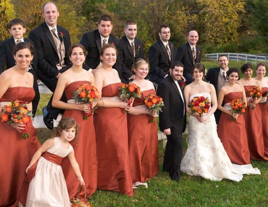 Wedding party in rust colored dresses and black suits with bride in white standing in front of white fence