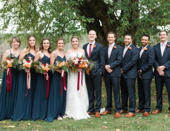 Wedding party in blue dresses and black suits with bride in white standing in front of large tree