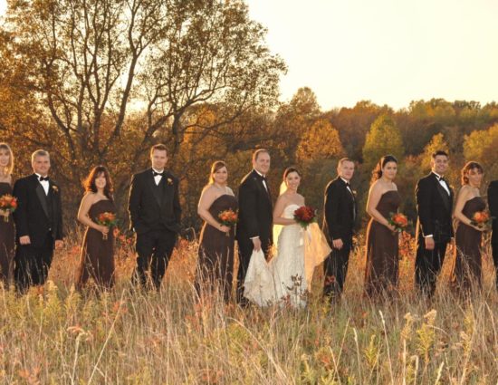 Wedding party in brown dresses and black suits with bride in white standing in a field of grasses with trees in the background with fall color