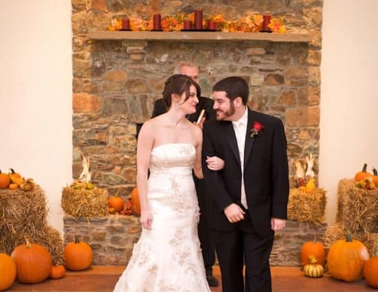 Bride in white dress and groom in black suit standing in front of stone fireplace decorated with bales of hay and orange pumpkins