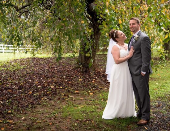 Bride in white dress and groom in gray suit standing under large trees with green leaves