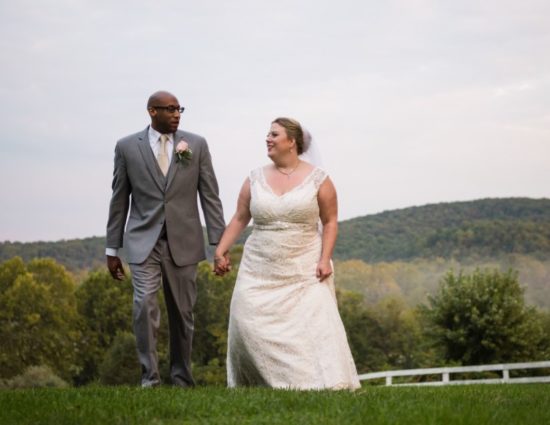 Bride in white dress and groom in gray suit standing in grass with rolling hills behind them