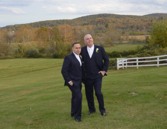 Two grooms in dark navy suits standing in green grass with rolling hills with fall color behind them