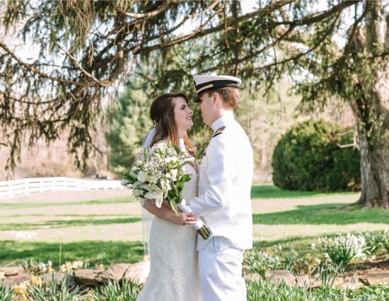 Bride in white dress and groom in white naval uniform standing on stone path surrounded by yellow and white flowers