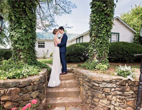 Bride in white dress and groom in dark blue suit standing on steps in front of white cottage