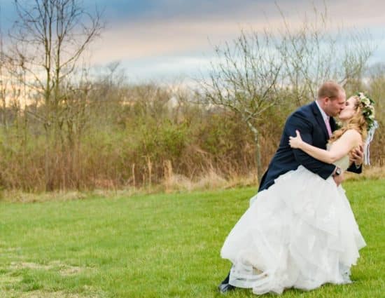 Bride in white dress and groom in dark blue suit standing in grass with sun setting behind them