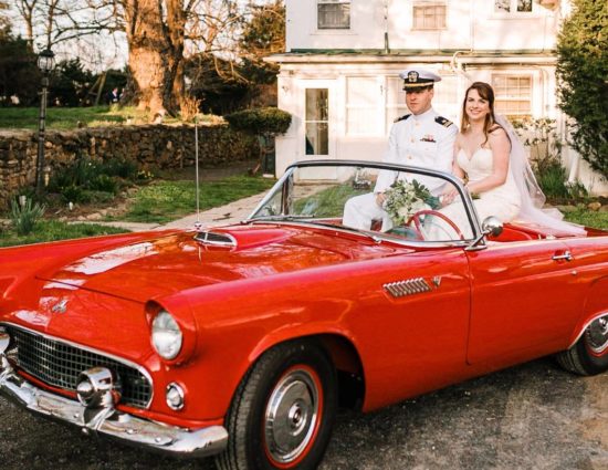 Bride in white dress and groom in white naval uniform sitting in old vintage red car