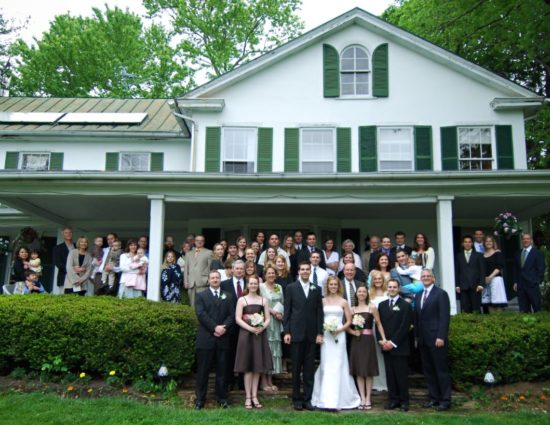 Entire wedding party with family posing in front of main house painted white with green shutters and roof