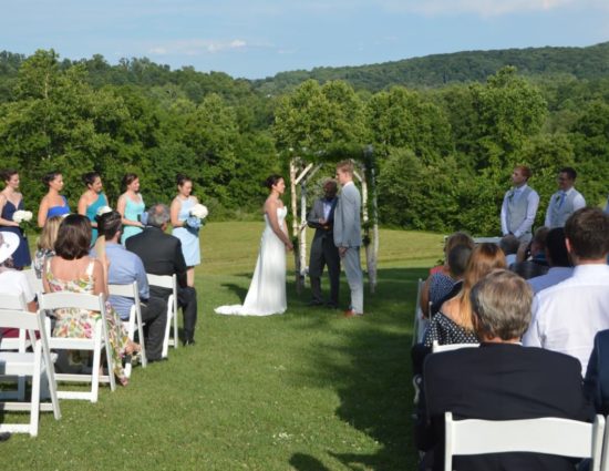 Wedding ceremony with bride in white dress and groom in light gray suit standing in front of wooden wedding alter with rolling hills in background