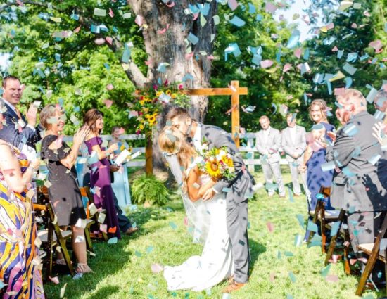 Bride in white dress and groom in gray suit kissing in the middle of the aisle with confetti falling around them