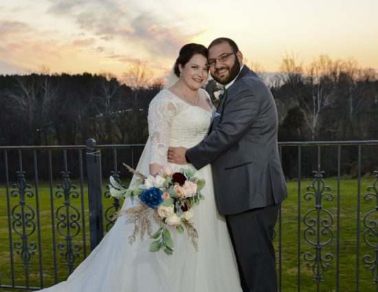 Bride in white dress and groom in gray suit standing by rod iron railing as sun sets behind