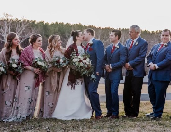 Wedding party with bridesmaids in light pink floral dresses, bride in white, and groom and groomsmen in blue suits