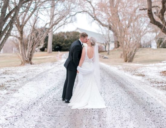 Bride in white dress and groom in black suit standing on gravel road covered in snow