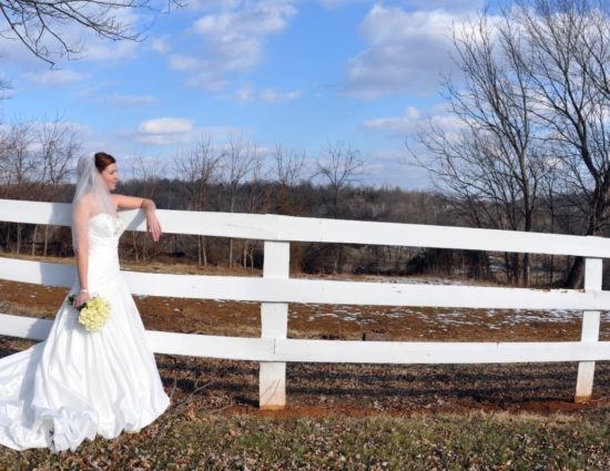 Bride in white dress standing by white fence holding bouquet of white roses