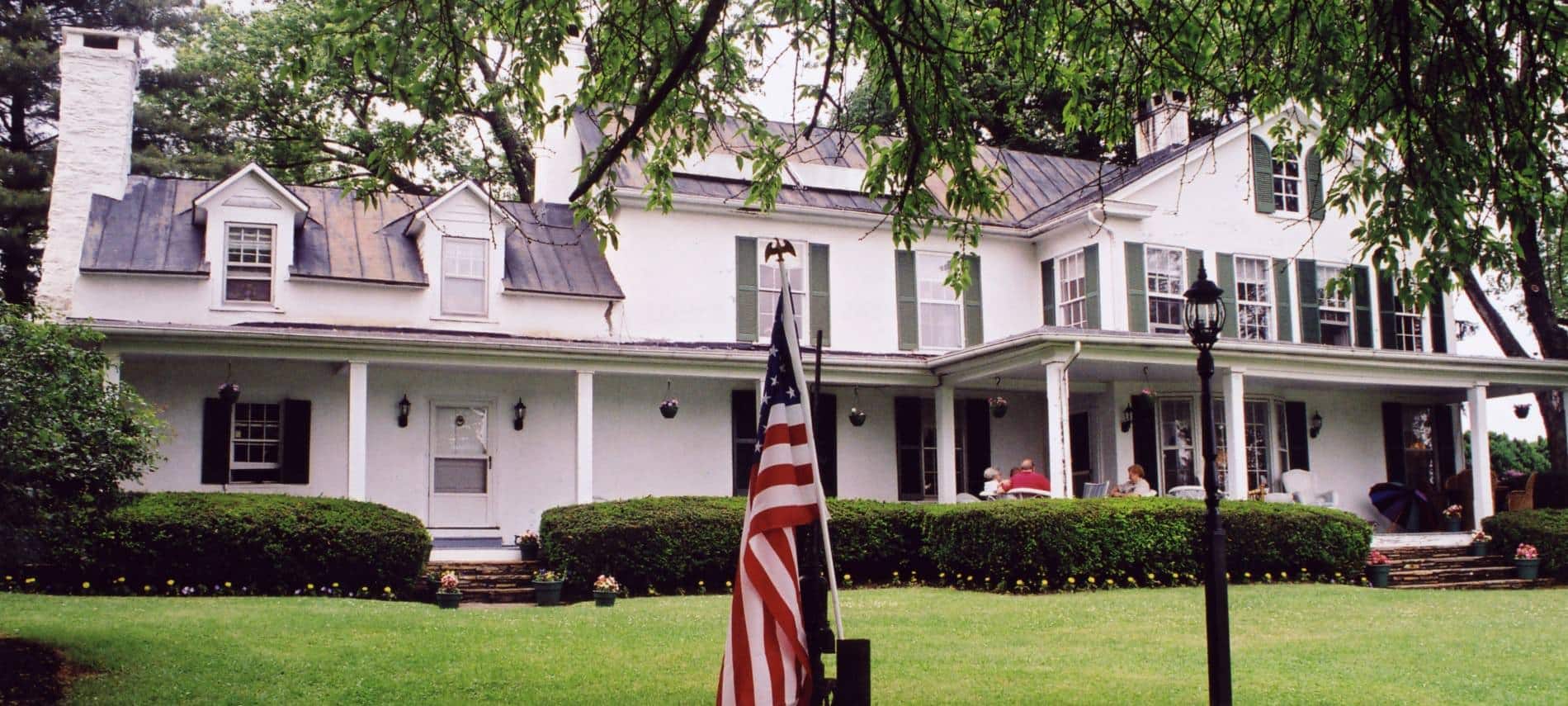 Exterior view of property painted white with dark shutters surrounded by a large green lawn and trees