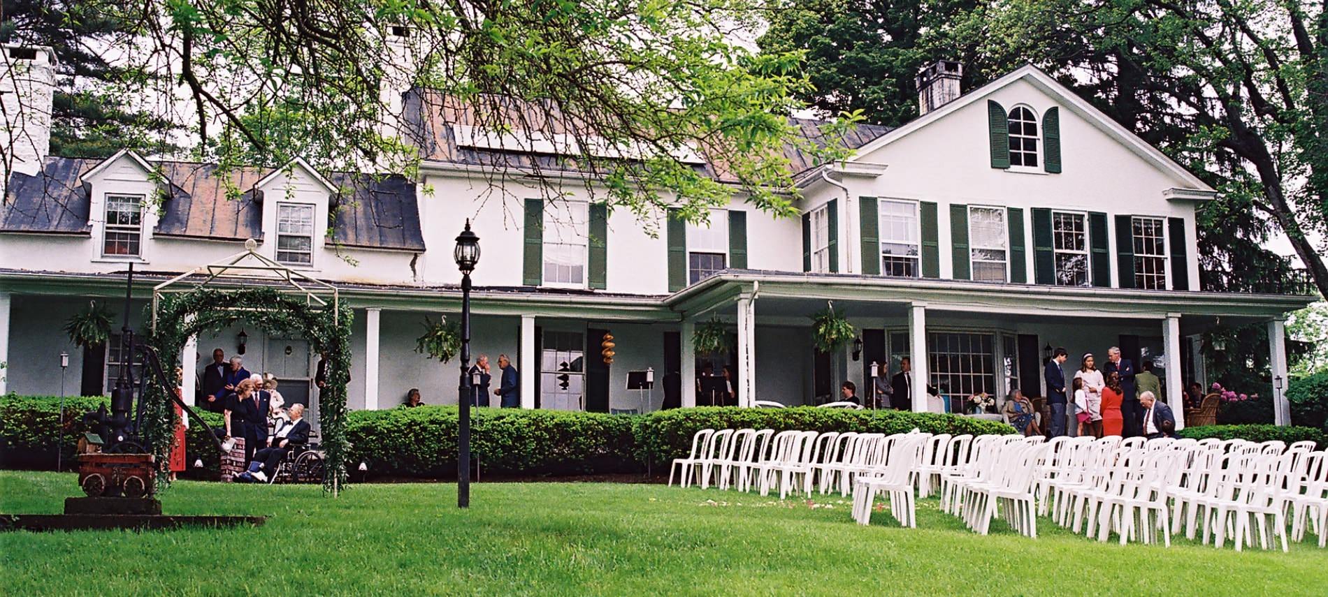 Exterior view of property painted white with dark shutters set up for a wedding with white chairs on the lawn