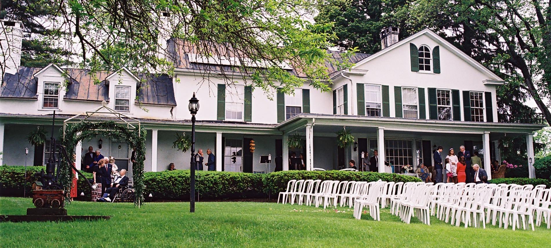 Exterior view of property painted white with dark shutters set up for a wedding with white chairs on the lawn