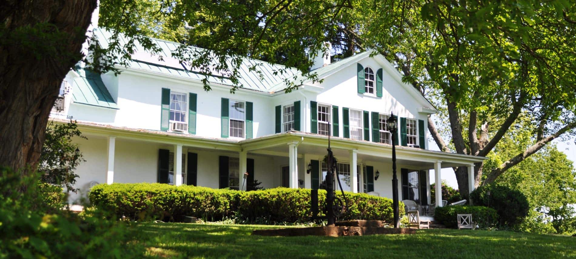Exterior view of property painted white with dark shutters surrounded by a large green lawn, shrubs, and trees