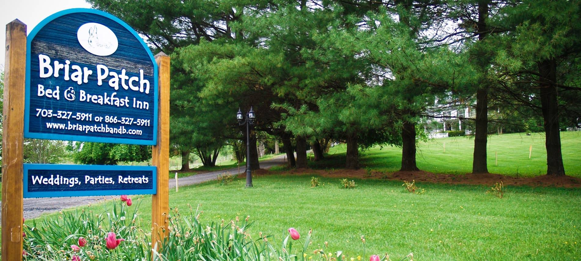 Large wooden sign painted blue with white lettering about Briar Patch Bed & Breakfast Inn surrounded by green grass, evergreen trees, and pink tulips