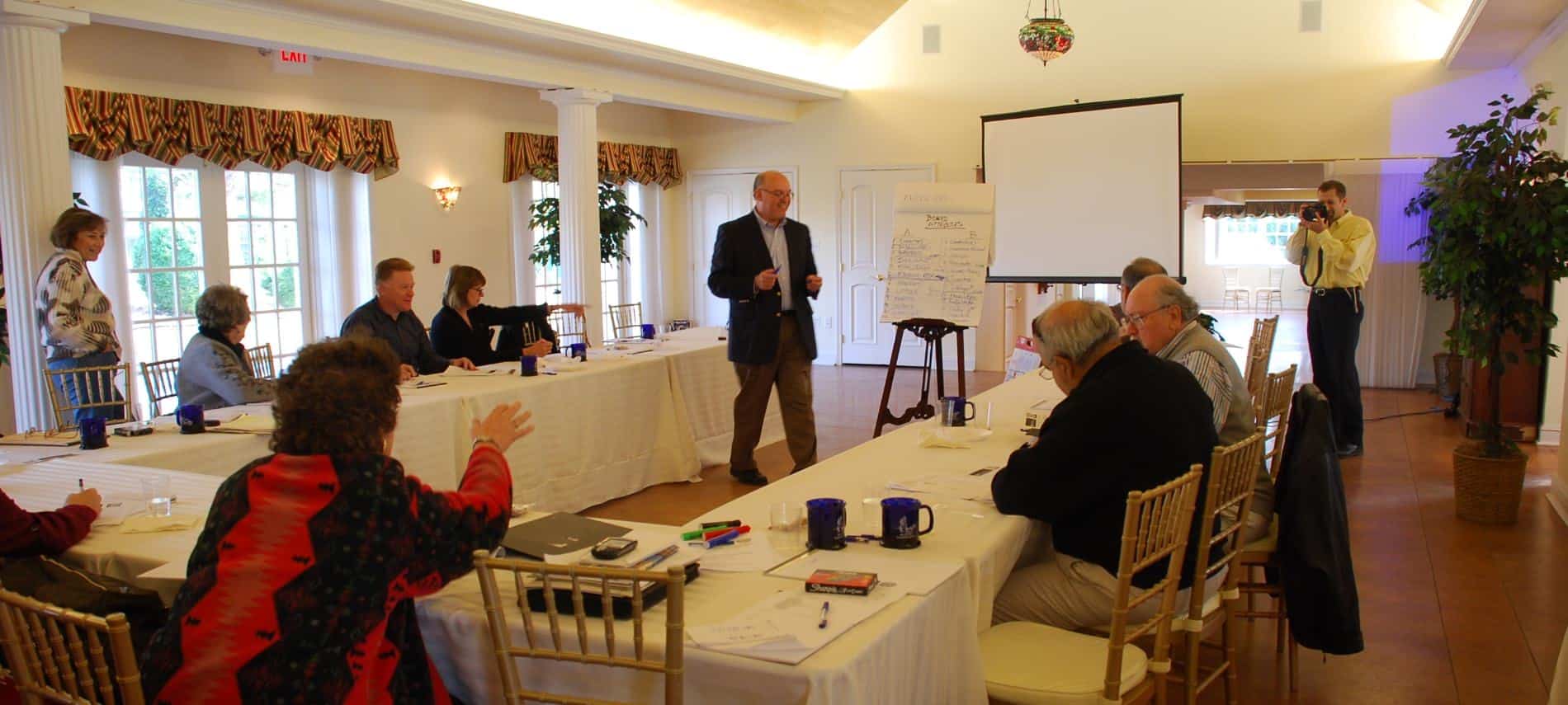 Large room set up with rectangle tables with white tableclothes in the shape of a U with people sitting in chairs listening to a presentation