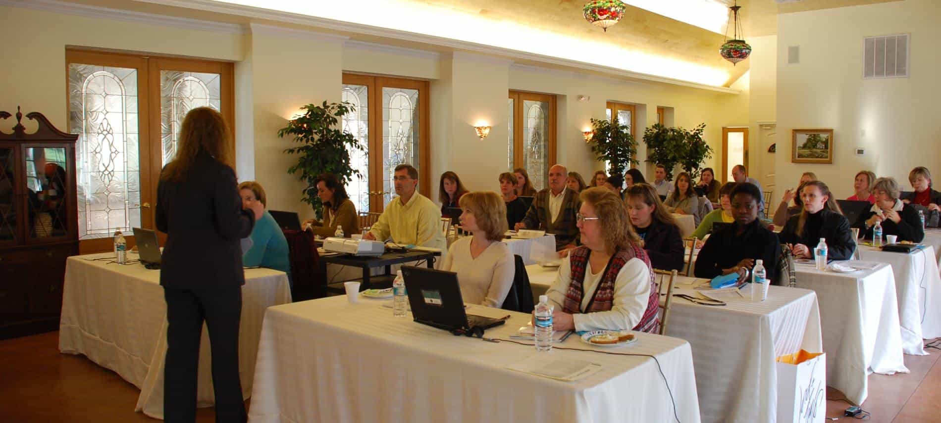 Large room set up with rectangle tables with white tableclothes with people sitting in chairs listening to a presentation