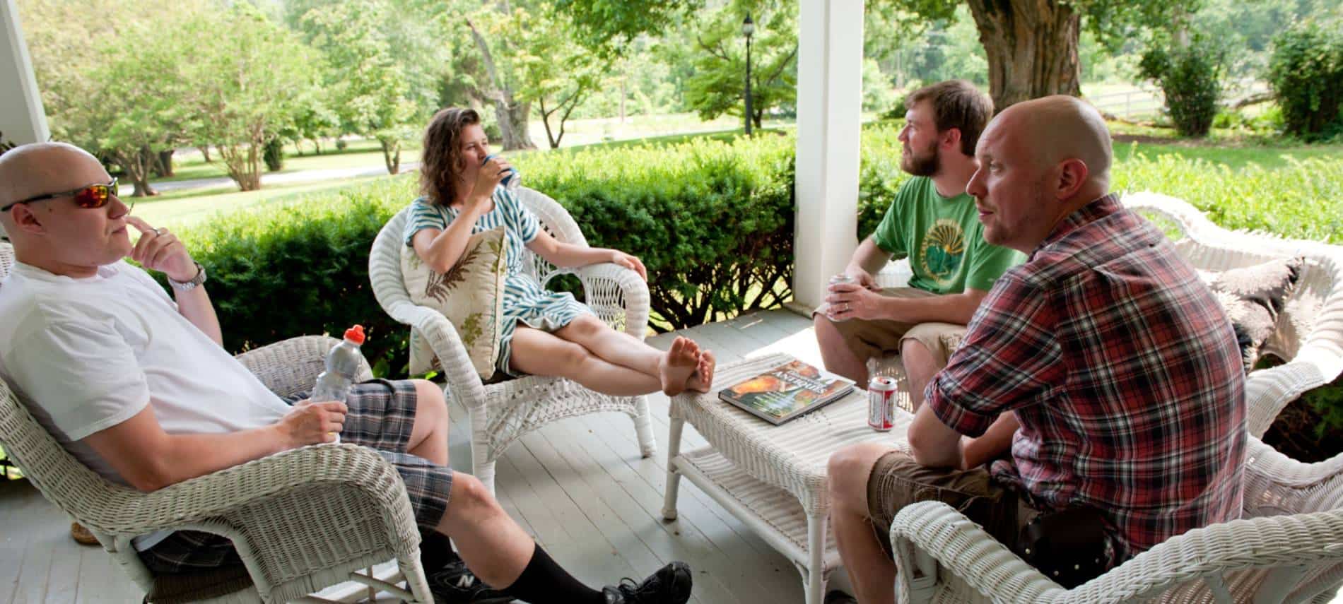 Four people sitting on white wicker patio furniture on front porch surrounded by green shrubs