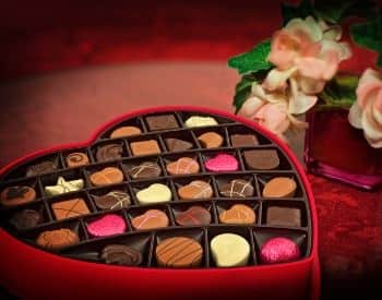 Large red valentine box filled with assorted chocolates next to pink vae with white and pink flowers