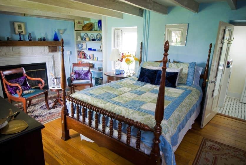 Bedroom with turquoise, sage green, and white bedding, light turquoise walls, hardwood flooring, fireplace with white stone and wooden mantel, and a sitting area