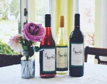 Three wine bottles, one red, one white, one rose, next to small glass vase with pink and purple flowers