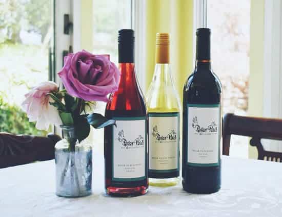 Three wine bottles, one red, one white, one rose, next to small glass vase with pink and purple flowers