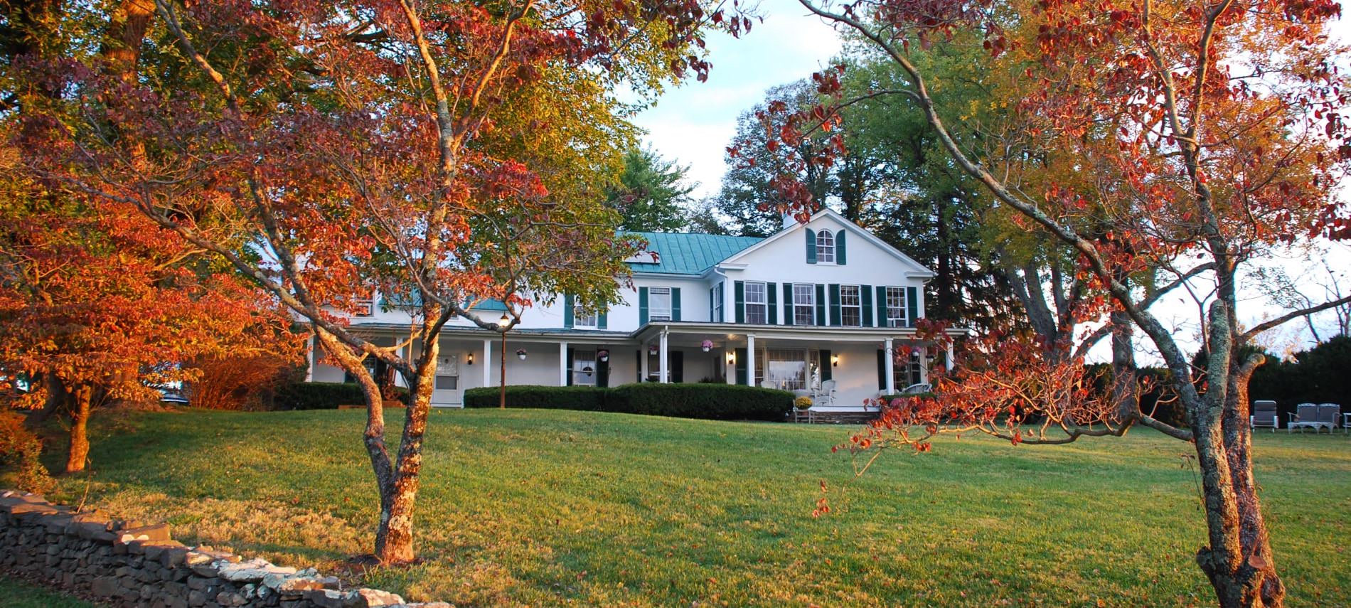 Exterior view of property painted white with dark shutters surrounded by a large green lawn and trees with fall color