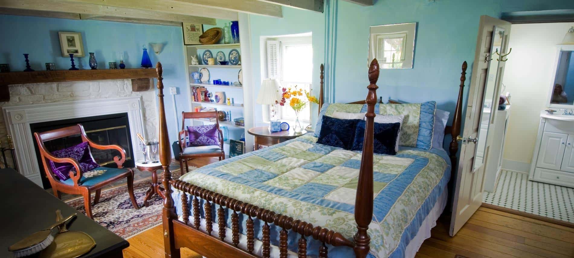 Bedroom with turquoise, sage green, and white bedding, light turquoise walls, hardwood flooring, fireplace with white stone and wooden mantel, and a sitting area