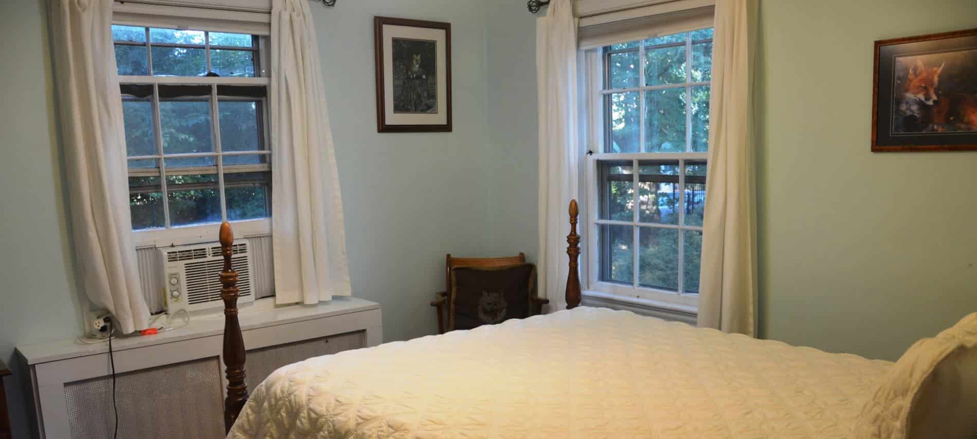 Bedroom with dark wooden four-post bed with cream quilted bedding, two windows with white trim and white curtains, and light blue walls