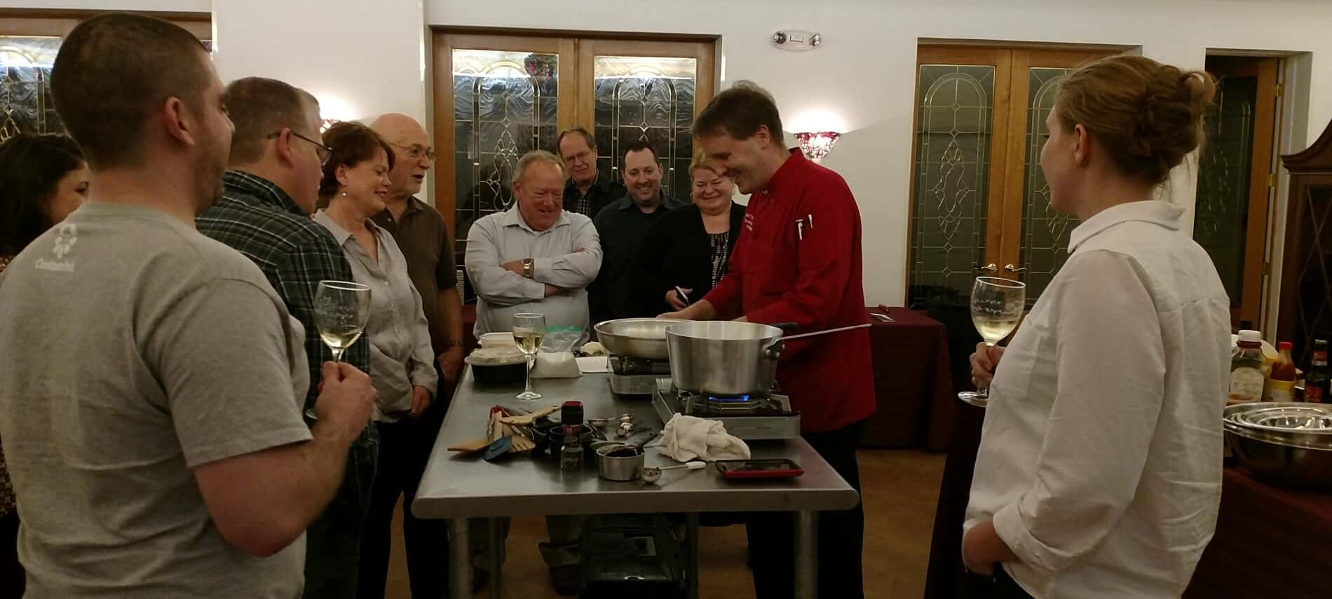 Smiling cooking class attendees standing around table in middle of cream room with smiling Chef Matt in red shirt demonstrating preparation of food.
