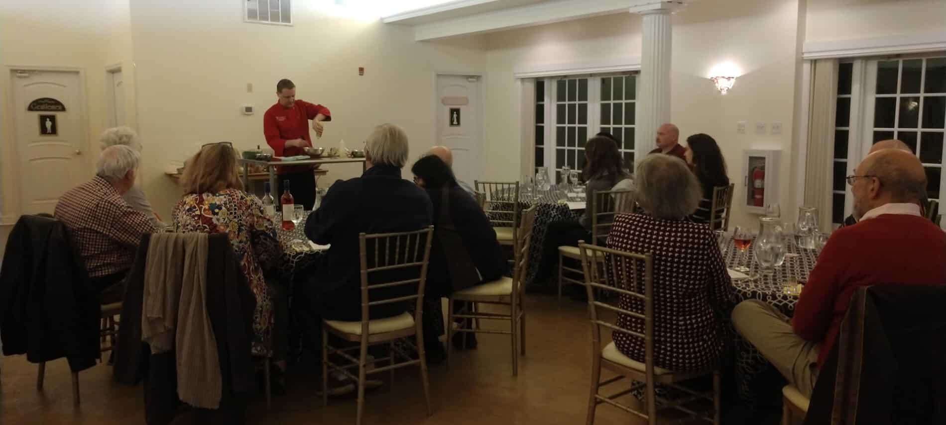 Group of cooking class attendees sitting a tables in dining area watching Chef Matt in red shirt demonstrate preparation of entree.