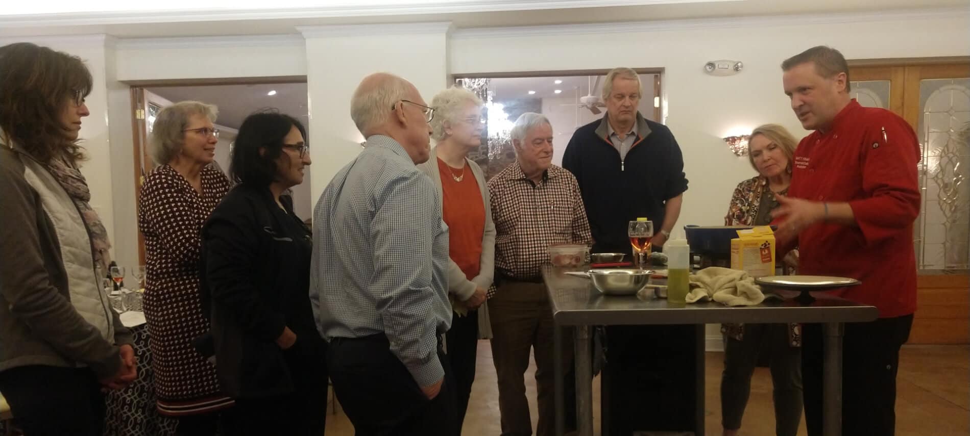 Group of cooking class attendees listening to Chef Matt in red shirt while standing around table in middle of cream room.